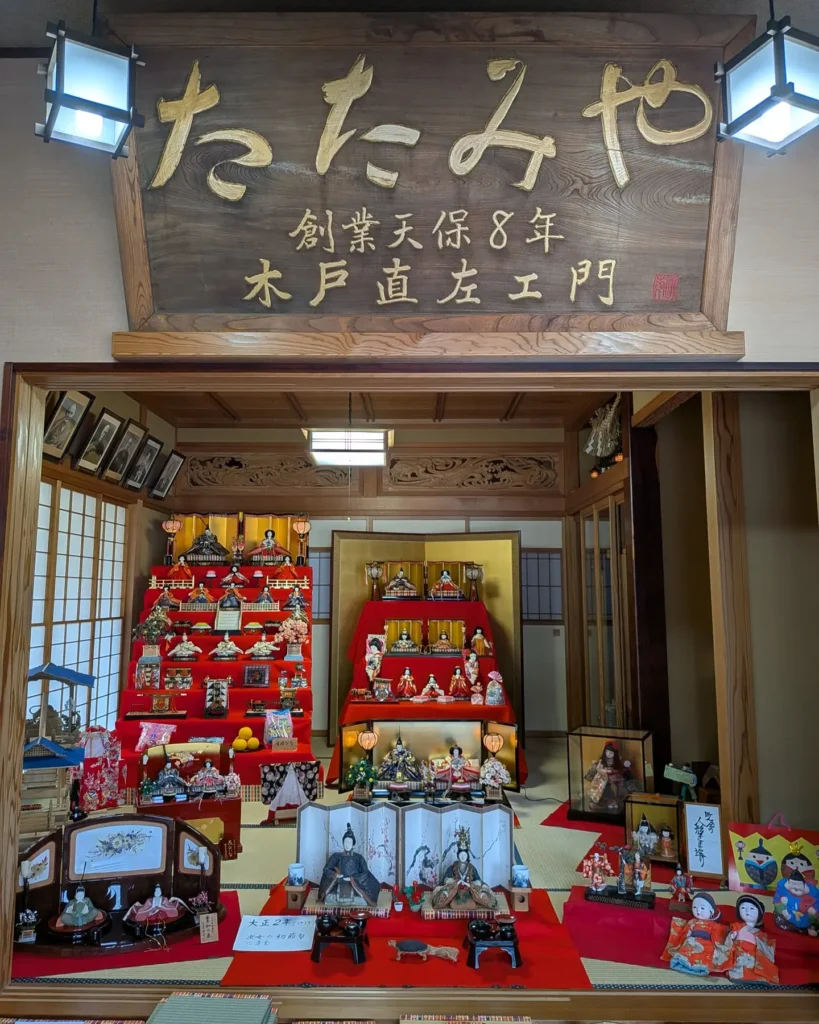 欅の一枚板による大きな看板のもと、緋毛氈の上に何組もの雛飾り