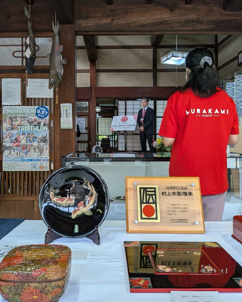 村上木彫堆朱の展示作品が並ぶ会場で挨拶する小杉和也代表理事